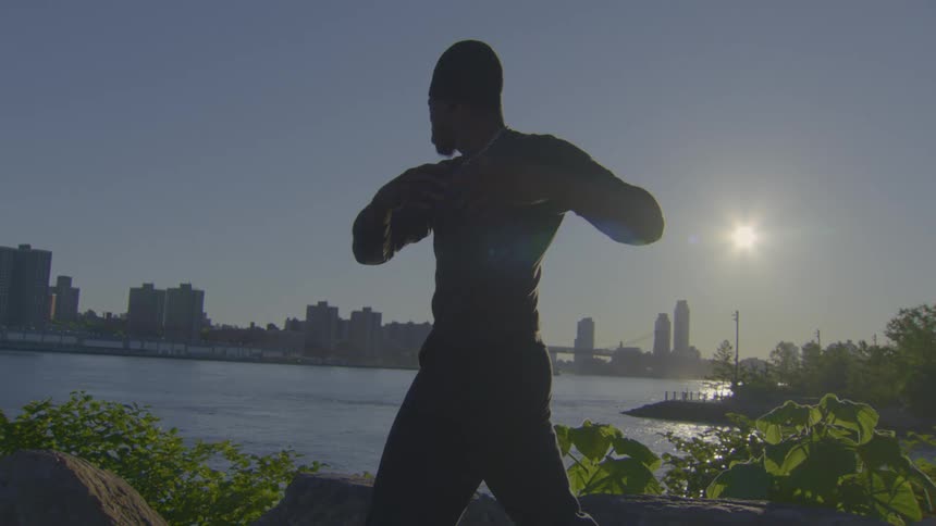 A silhouetted man performs slow, meditative Tai Chi movements by the river as the sun rises over the city skyline