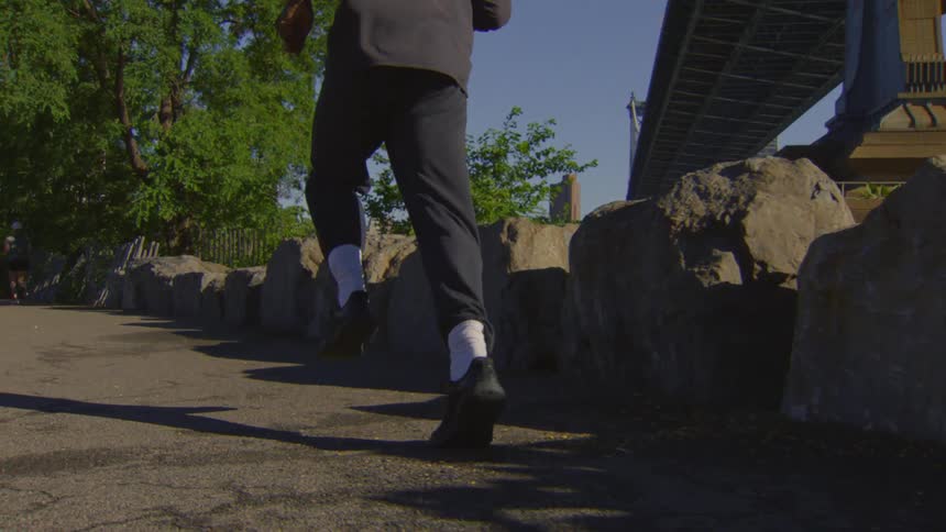 Slow motion low angle shot follows the legs and feet of a person jogging on a paved path under a bridge in a city park