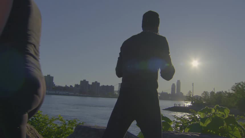 A silhouetted man performs slow, graceful tai chi movements by a river as people walk by with the city skyline in the background