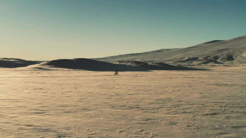 A lone cyclist pedals a fat bike across a vast, windswept, and sunlit snowy landscape