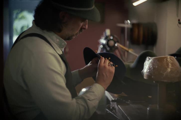 An artisan hat maker carefully hand-stitching a new hat in his dimly lit workshop
