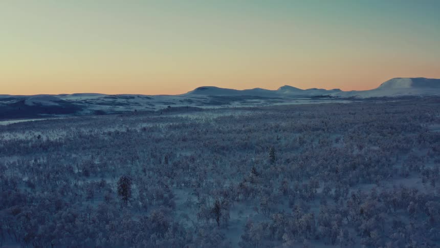 An expansive aerial view slowly pulls back over a vast, snow-covered forest wilderness as the sun sets over distant mountains