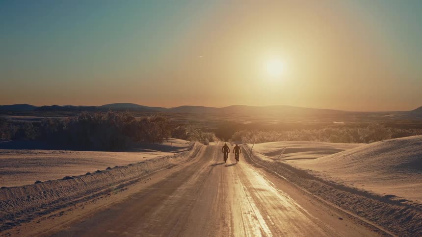 Two cyclists ride their bikes down a long, scenic road in a vast, snowy landscape at sunset