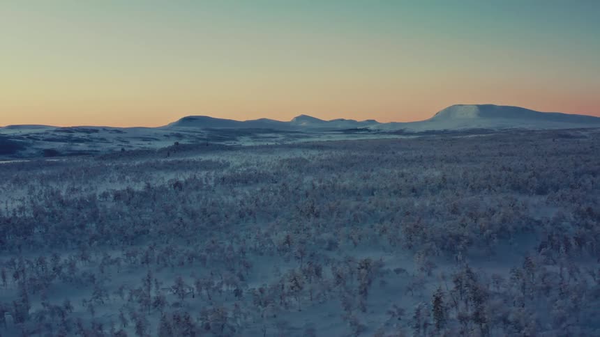 An aerial view pulls back over a vast, snow-covered forest tundra with distant mountains glowing in the serene light of dusk