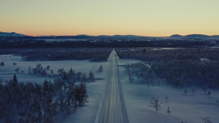 A serene aerial view follows a long, straight road through a vast, snow-covered arctic landscape during a beautiful winter sunset
