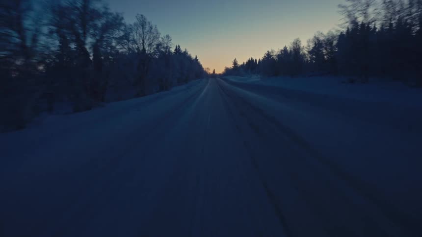 Driving along a desolate, snow-covered road through a frosted forest landscape at dusk