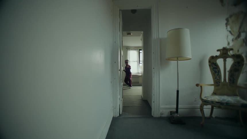 A slow-moving camera shot travels down a dark, rundown hallway towards a mysterious masked figure practicing karate alone in an empty room