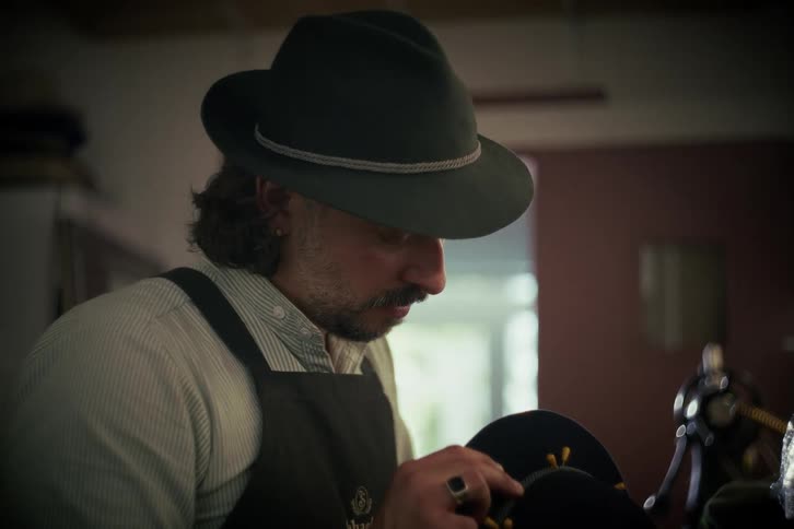 A focused artisan hat maker meticulously hand-sews the final details of a traditional hat in his workshop