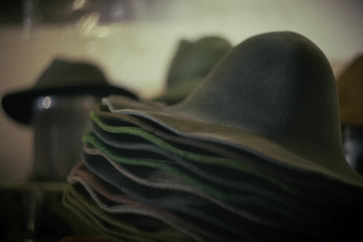 A stack of colorful, unfinished felt hats sits on a shelf in a dimly lit workshop