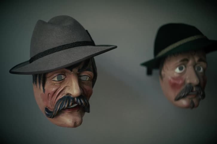 Two eerie traditional German carnival masks with hats and mustaches hang on a wall in moody lighting