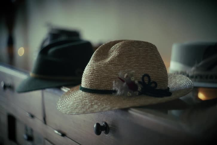A slow focus pull reveals a collection of traditional hats, including a straw fedora and a green Tyrolean hat, resting on a vintage wooden cabinet