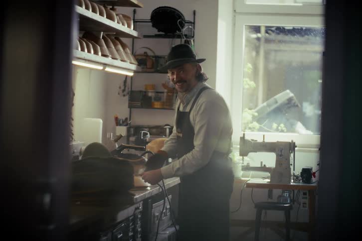A cheerful hatter with a mustache puts on his gloves and starts working on a new hat in his cozy, sunlit workshop
