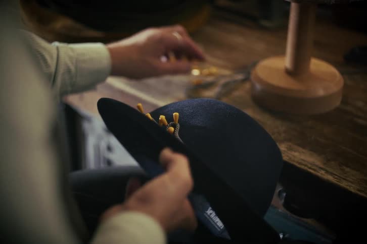 A skilled artisan meticulously pins a decorative cord onto a custom-made felt hat in a traditional workshop