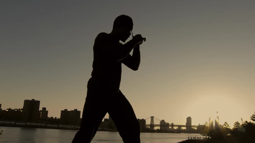Silhouette of a man practicing boxing punches in slow motion by a river with a city skyline in the background during sunset