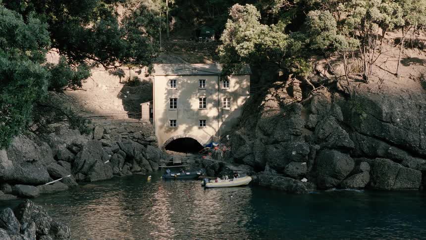 A picturesque old boathouse is tucked away in a tranquil, rocky cove on a sunlit day, with small boats floating on the calm sea