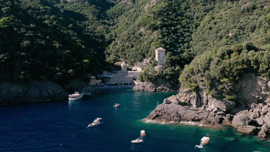 A slow aerial approach reveals the breathtaking San Fruttuoso Abbey and Doria Tower nestled in a secluded cove with boats on the Portofino coast