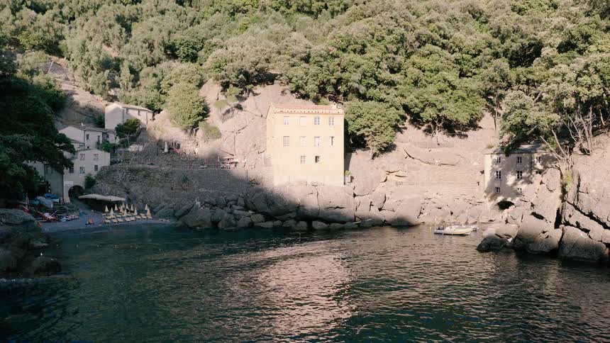 Aerial view pushing in on a scenic secluded cove on the Italian coast, where historic buildings are built into the rocky, tree-lined cliffs
