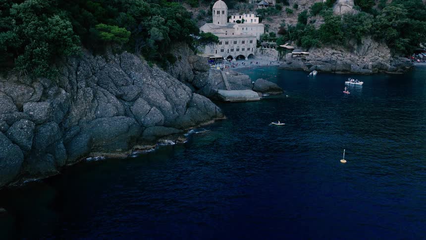 An aerial drone flies over the deep blue sea to reveal the historic San Fruttuoso Abbey and beach cove with swimmers and boats