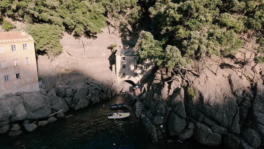 A static high-angle aerial shot captures a picturesque hidden cove with old stone buildings nestled into the rocky, tree-lined cliffs as small boats float in the water
