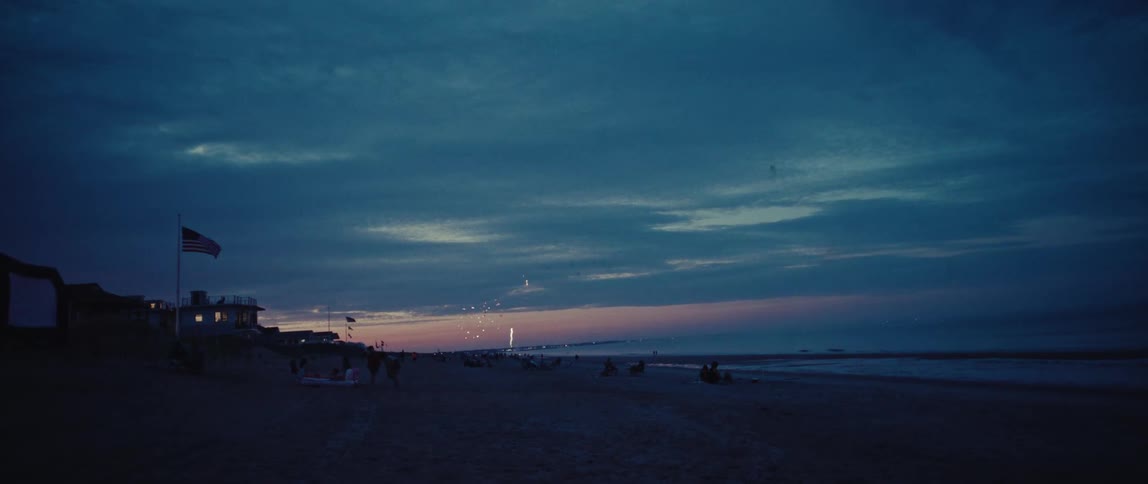 A firework explodes in the dusky sky over a beach with an American flag waving in the foreground