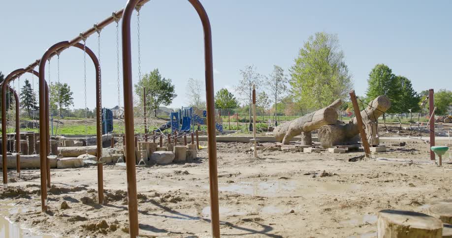 A slow dolly shot reveals an empty playground with swings and logs under construction in a muddy park
