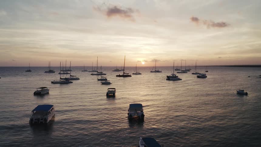 An aerial view slowly descends over numerous boats and catamarans anchored in the calm ocean during a beautiful golden sunset