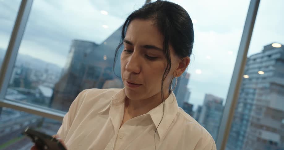 A concerned businesswoman reacts to bad news on her smartphone in a modern high-rise office with a city view