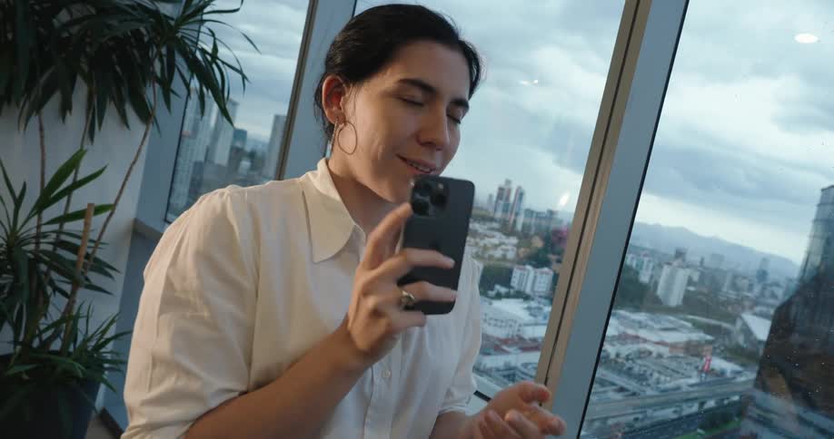 A happy businesswoman in a modern office smiles thoughtfully after using her smartphone, with a stunning city view in the background