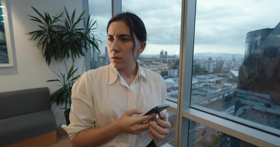 A businesswoman's expression shifts from concern to a happy smile while using her phone by a window overlooking the city