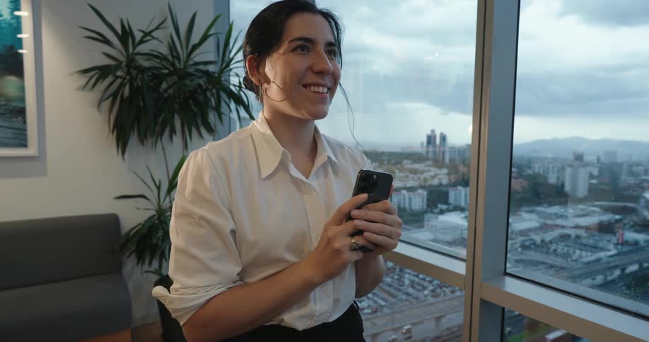 An ecstatic businesswoman joyfully communicates on her smartphone, beaming with happiness in her high-rise office overlooking the cityscape