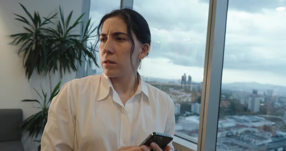 A young businesswoman stands by her office window, her expression shifting from concern to a joyful smile after checking her phone