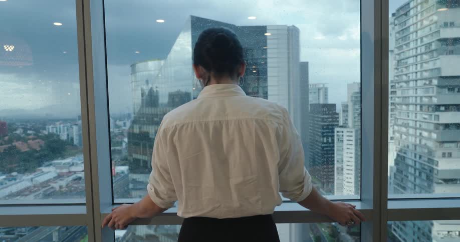A businesswoman in a white shirt contemplates the sprawling cityscape from her high-rise office window on a rainy day
