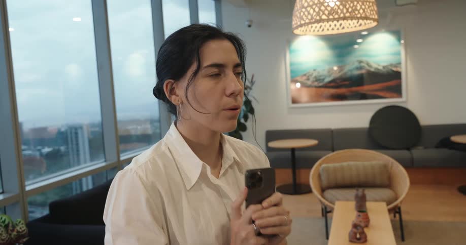 A focused businesswoman walking in a modern office with city views stops to take a deep, relaxing breath while holding her smartphone