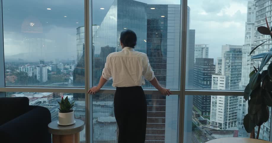 A pensive businesswoman approaches the large window in her high-rise office to look out at the sprawling city on an overcast day