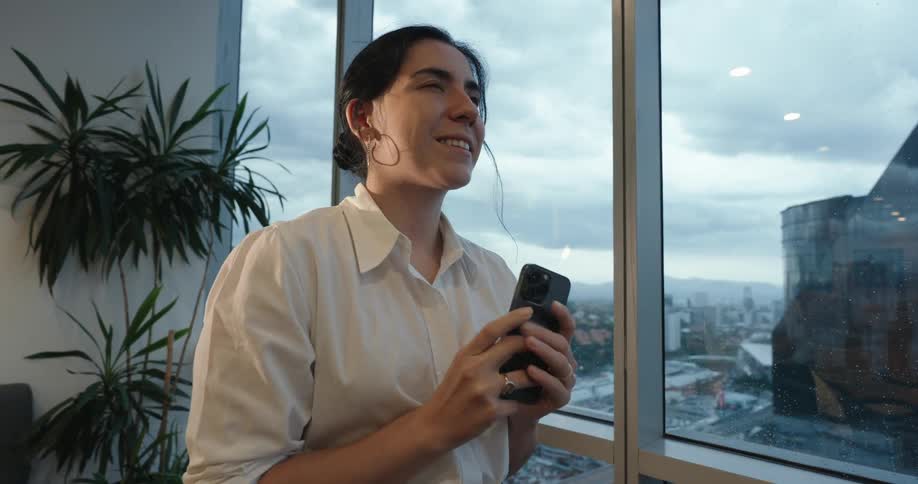 An elegant businesswoman smiles with joy as she listens to music on her phone while looking out the window of a high-rise office