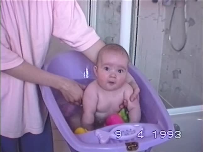 A vintage home video from 1993 shows a mother bathing her baby in a small purple tub while the child splashes and plays with toys