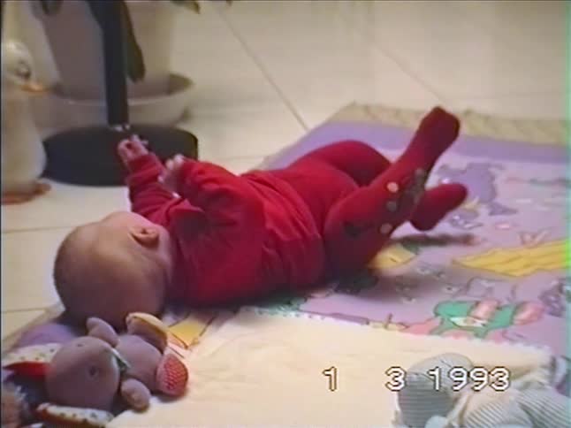 A nostalgic 1993 home video shows an infant in a red onesie getting fussy during tummy time and rolling onto its back