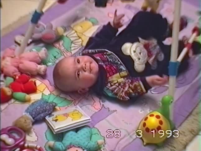 A sweet baby smiles for the camera while lying on a colorful playmat in a nostalgic home video from 1993