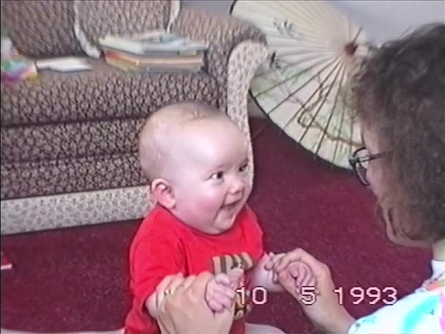 An adorable baby sitting on a lap bursts into joyful laughter while playing a hand game in this authentic 1993 home movie