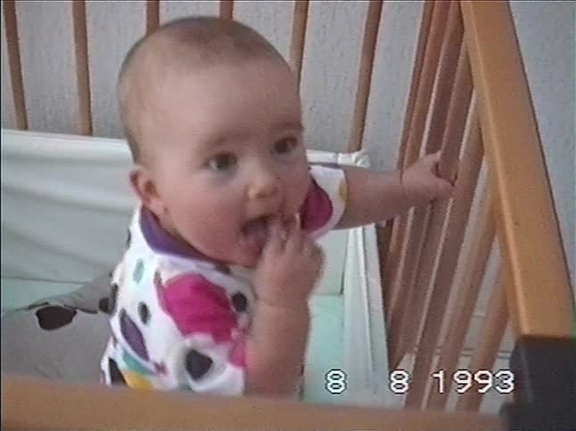 A vintage home video from 1993 shows a cute baby in a colorful onesie smiling happily while standing in a wooden crib