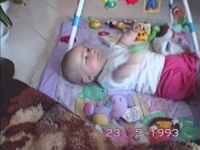 A cute baby lies on a colorful playmat, laughing and playing with toys in an authentic home video from 1993