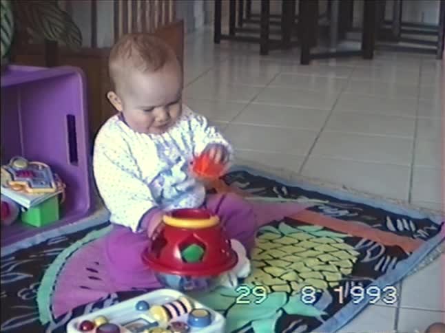 An adorable baby sits on a colorful playmat and intently plays with a shape-sorter toy in this vintage 1993 home video