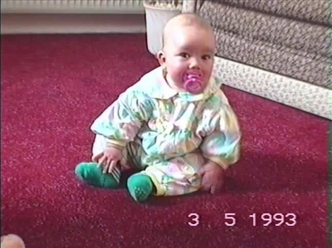 A cute baby in a colorful onesie sits on a red carpet before slowly rolling over onto its back in this 1993 home video