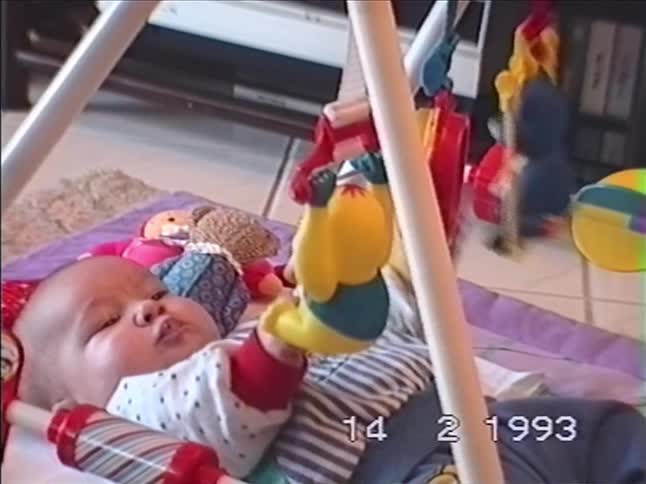 A newborn baby lies on a playmat, curiously looking at the colorful toys hanging from a baby gym in 1993