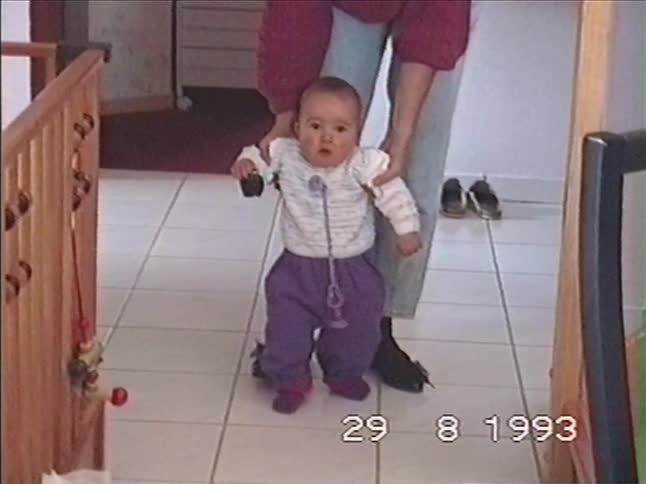 A parent helps their wobbly baby learn to walk at home in a cute home video from 1993