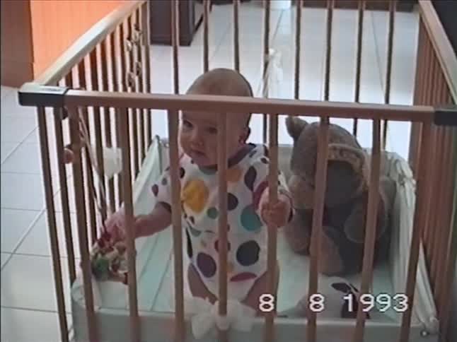 A cute baby in a colorful polka-dot onesie smiles happily while standing in a wooden playpen in this 1993 home video