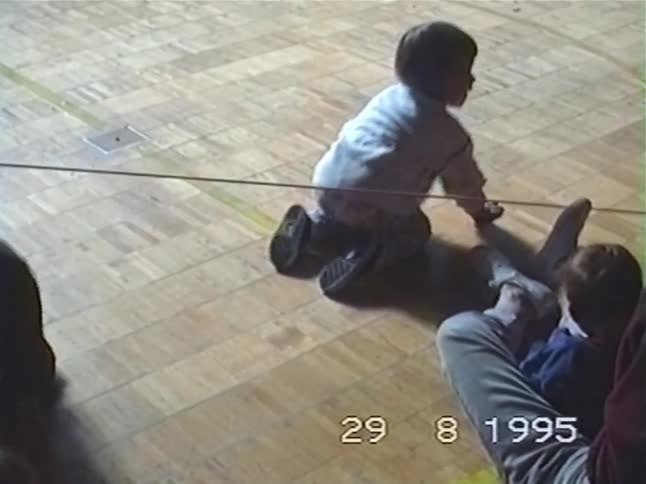 A vintage 1995 home video shows toddlers crawling and playing together on a wooden gymnasium floor