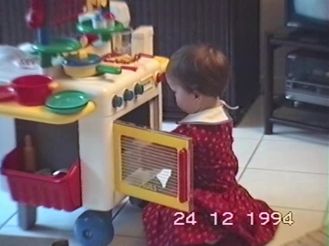 Adorable toddler in a festive red dress explores her new toy kitchen in a nostalgic home video from 1994