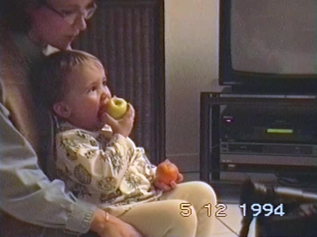 A cute toddler eats an apple while sitting on their mother's lap and watching TV in this authentic 1994 home video
