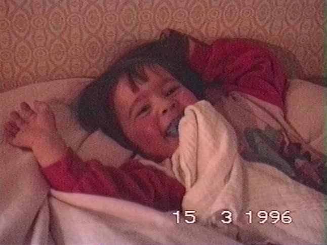 Adorable toddler with a pacifier and rosy cheeks wakes up in bed and smiles happily in a vintage home video from 1996